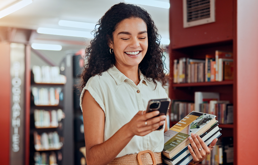 Mujer joven con cabello rizado en una biblioteca, sonriendo y mirando su teléfono, con libros en las manos.