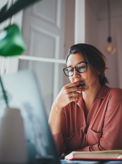 Mujer joven con gafas y cabello recogido, sentada en un escritorio. Mira fijamente una laptop mientras sostiene un bolígrafo cerca de su mentón, en una pose reflexiva