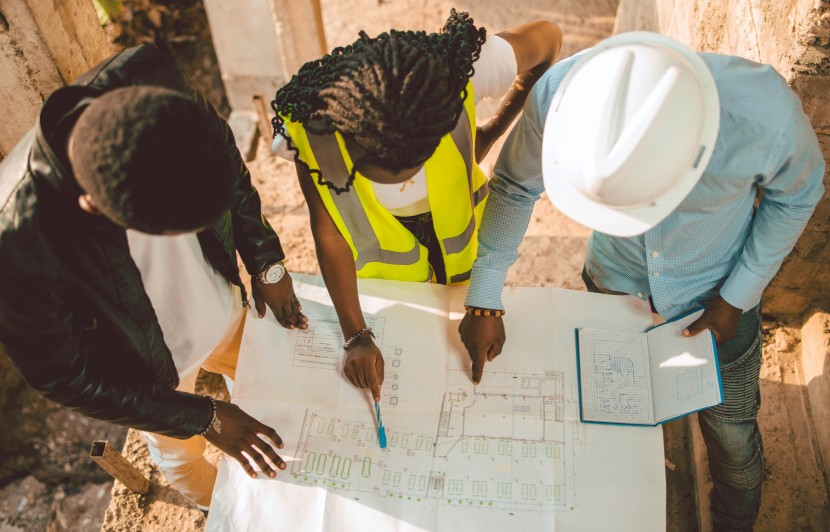 Equipo de ingenieros trabajando y revisando planos arquitectónicos en una obra de construcción, con casco de seguridad y chalecos reflectantes.