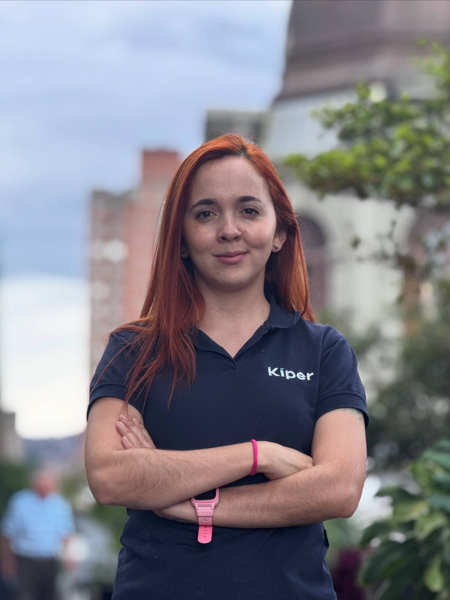 Una mujer joven de pie al aire libre, con una postura segura y los brazos cruzados. Tiene el cabello rojizo y lacio, y está usando una camiseta tipo polo negra con el logo “Kiper” en el pecho. Lleva un reloj o pulsera inteligente rosada en la muñeca. Su e