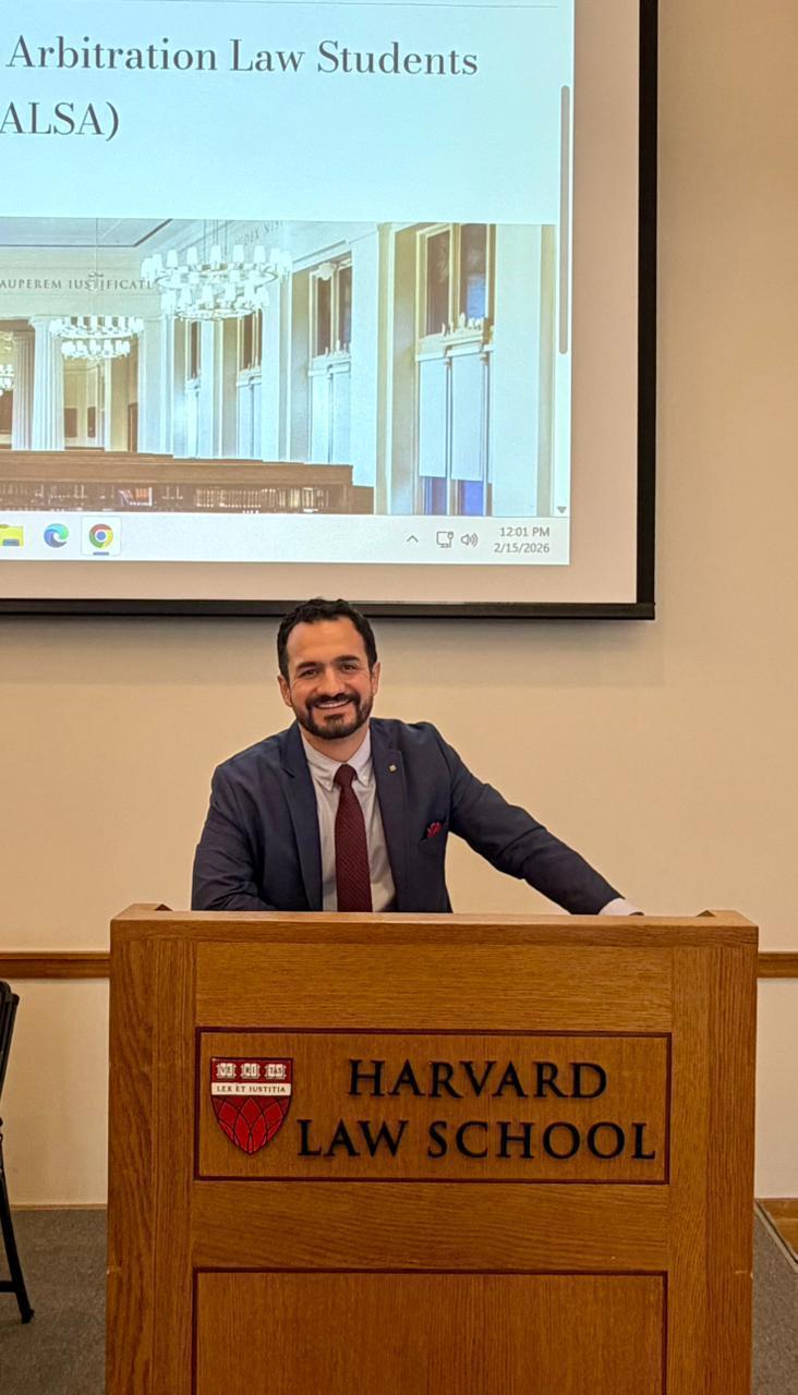 Retrato de medio cuerpo del profesor Alirio Giraldo sonriendo en el podio de la Harvard Law School, con la pantalla de proyección visible al fondo