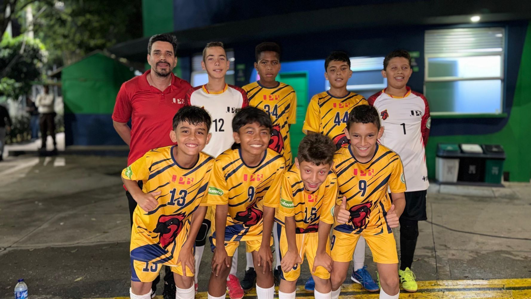 Un equipo infantil de fútbol posa sonriente junto a su entrenador por la noche, vistiendo uniformes amarillos con azul de la UPB y uniformes blancos de portero.
